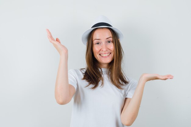 Junge Dame, die Schuppengeste im weißen T-Shirt-Hut macht und fröhlich aussieht