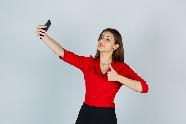 Junge Dame, die selfie nimmt, während Daumen in roter Bluse, Rock zeigt und charmant aussieht