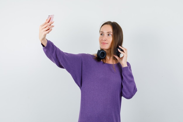 Junge Dame, die selfie nimmt, während sie Kopfhörer im violetten Hemd, Vorderansicht hält.