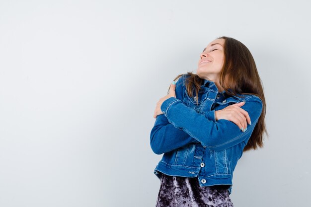 Junge Dame, die sich in Bluse, Jeansjacke umarmt und entspannt aussieht, Vorderansicht.