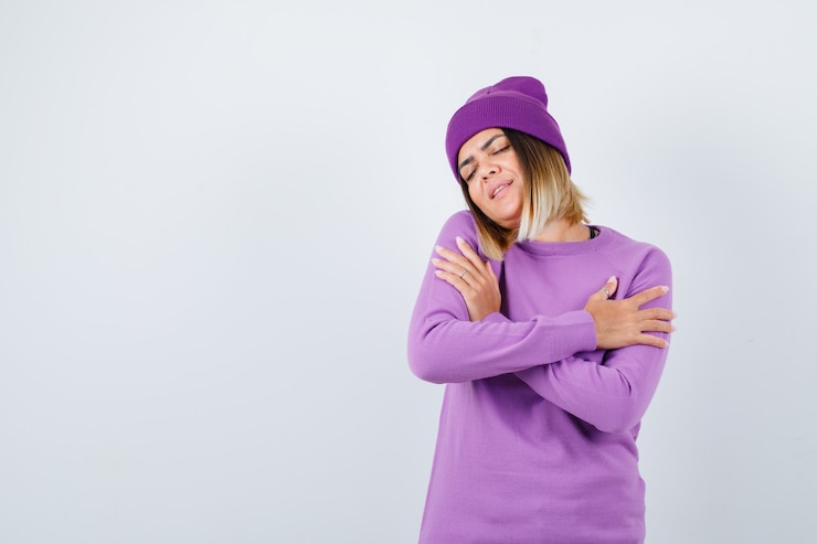 Kostenlose Foto junge dame, die sich in lila pullover, mütze umarmt und friedlich aussieht. vorderansicht.