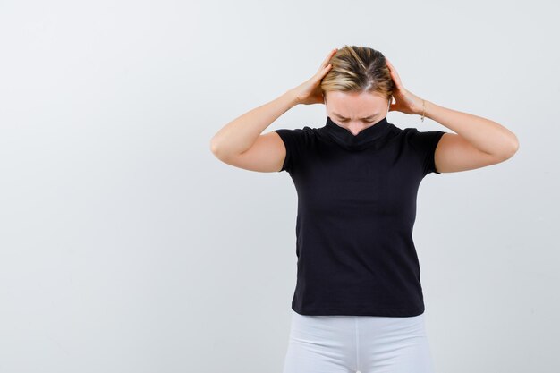 Junge Dame, die sich in T-Shirt, Hose, medizinischer Maske die Hände am Kopf hält und müde aussieht