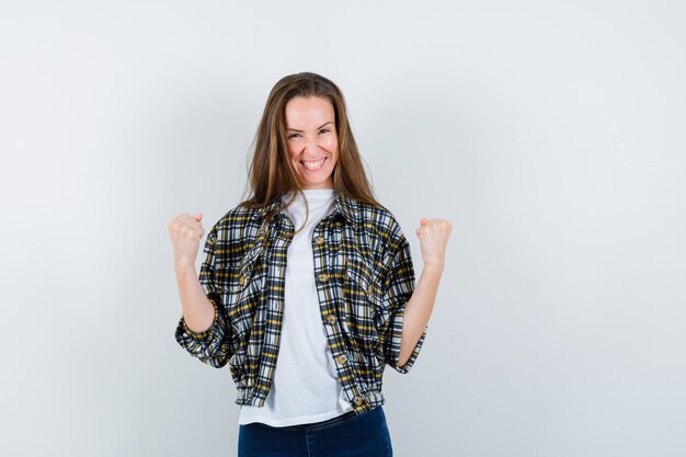 Junge Dame, die Siegergeste im T-Shirt, in der Jacke, in den Jeans zeigt und glückselig aussieht, Vorderansicht.