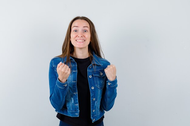 Junge Dame, die Siegergeste in Bluse, Jacke zeigt und glücklich aussieht. Vorderansicht.