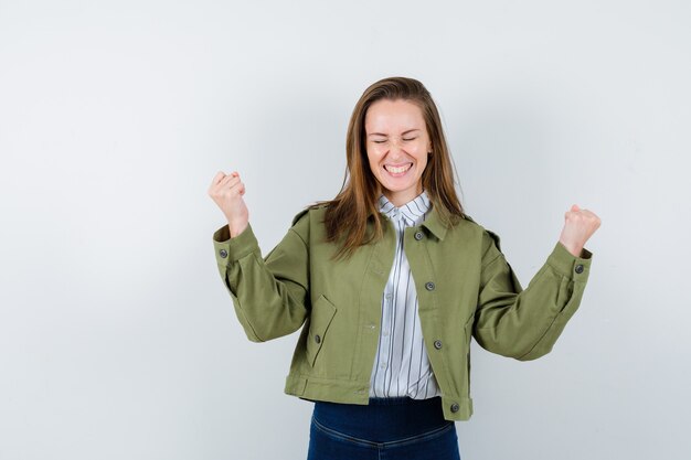 Junge Dame, die Siegergeste in Hemd, Jacke zeigt und glücklich aussieht, Vorderansicht.