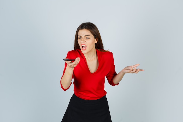 Junge Dame, die Sprachnachricht auf Handy in roter Bluse, Rock aufzeichnet und wütend schaut