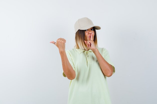 Junge Dame, die Stille Geste zeigt, während sie auf die linke Seite mit Daumen im T-Shirt, Mütze zeigt und selbstbewusst aussieht, Vorderansicht.