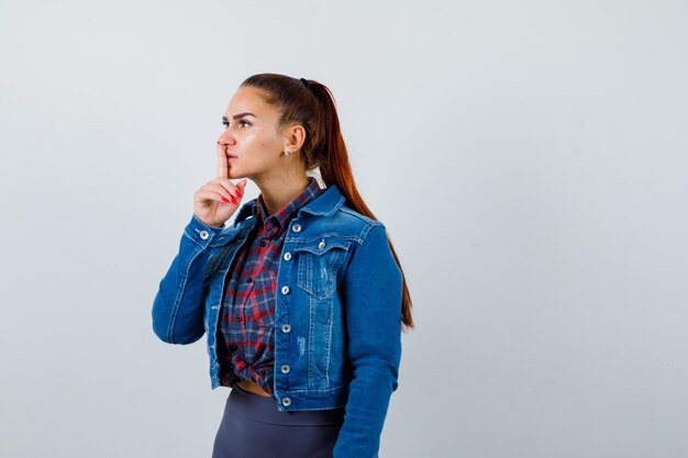 Junge Dame, die Stillegeste in kariertem Hemd, Jeansjacke zeigt und vorsichtig aussieht. Vorderansicht.