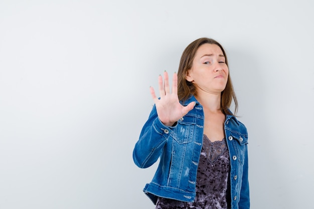 Junge Dame, die Stoppgeste in Bluse, Jeansjacke zeigt und ängstlich aussieht. Vorderansicht.