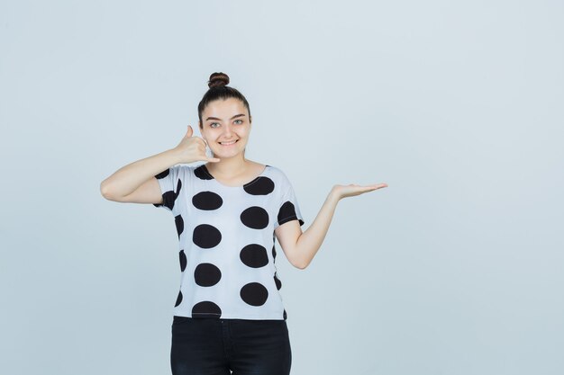 Junge Dame, die Telefongeste zeigt, während sie vorgibt, etwas in T-Shirt, Jeans zu halten und glücklich auszusehen. Vorderansicht.