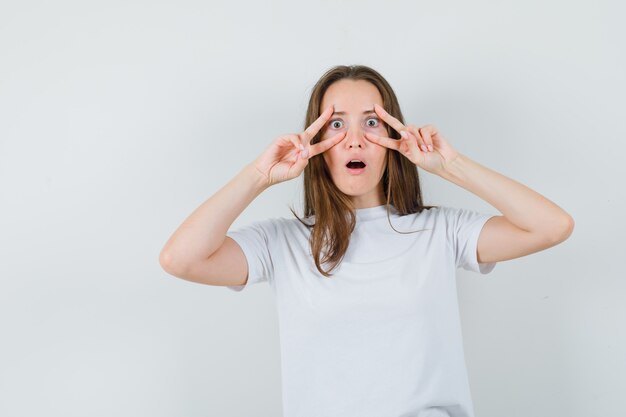 Junge Dame, die V-Zeichen auf Augen im weißen T-Shirt zeigt und erstaunt schaut