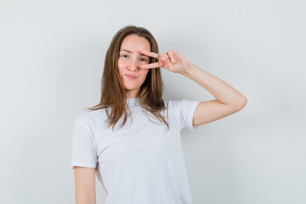 Junge Dame, die V-Zeichen nahe Auge im weißen T-Shirt zeigt und zuversichtlich schaut