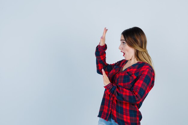 Junge Dame, die versucht, sich mit den Händen in einem karierten Hemd zu blockieren und verängstigt auszusehen. Vorderansicht.