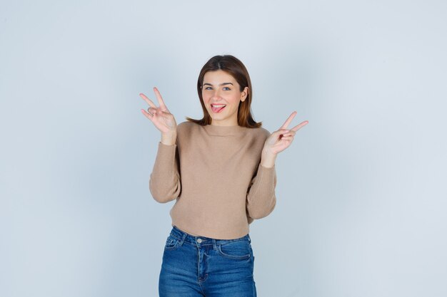 Junge Dame, die Victory-Zeichen in beigem Pullover, Jeans zeigt und fröhlich aussieht. Vorderansicht.