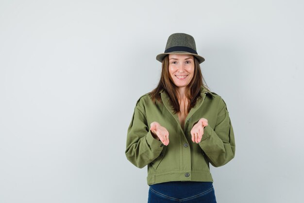 Junge Dame, die vorgibt, etwas in einem Jackenhosenhut anzubieten oder zu zeigen, und fröhlich aussieht