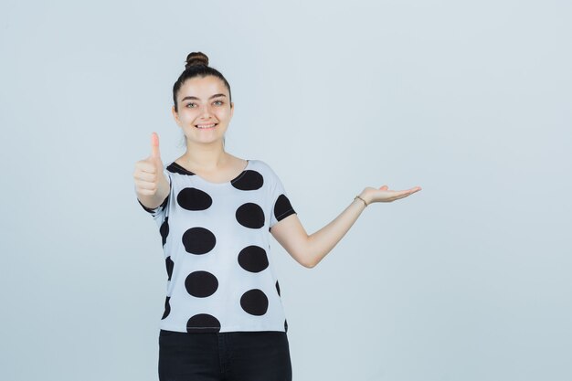 Junge Dame, die vorgibt, etwas zu zeigen, während Daumen in T-Shirt, Jeans zeigt und glückliche Vorderansicht schaut.