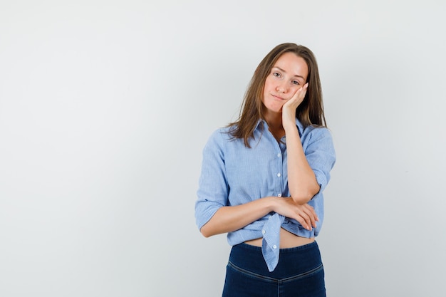 Junge Dame, die Wange auf erhabener Handfläche im blauen Hemd, in der Hose lehnt und nachdenklich schaut.