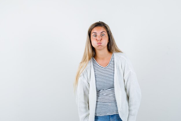 Junge Dame, die Wangen im T-Shirt, in der Jacke bläst und verwirrt, Vorderansicht schaut.