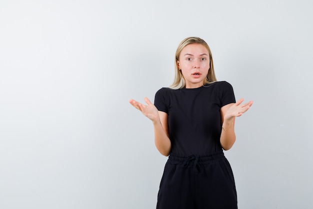 Junge dame, die zweifelgeste in t-shirt, hose zeigt und ängstlich aussieht. vorderansicht.