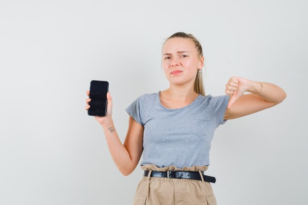 Junge Dame hält Handy, zeigt Daumen nach unten in T-Shirt und Hose und sieht unzufrieden aus