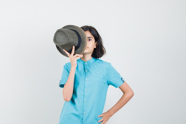 Junge Dame im blauen Hemd, das Hut auf ihrer Gesichtshälfte hält und seltsam aussieht