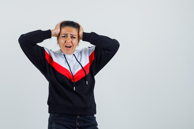 Junge Dame im bunten Sweatshirt, das Hände auf Kopf hält und alarmiert, Vorderansicht schaut.