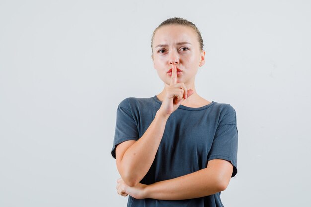 Junge Dame im grauen T-Shirt, das Schweigengeste zeigt und vorsichtig schaut