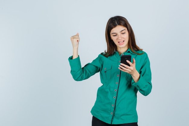 Junge Dame im grünen Hemd, das Handy betrachtet, Siegergeste zeigt und glückliches, Vorderansicht schauend.