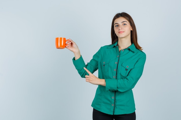 Junge Dame im Hemd, die orange Tasse Tee hält, auf obere linke Ecke zeigend und selbstbewusst, Vorderansicht schauend.