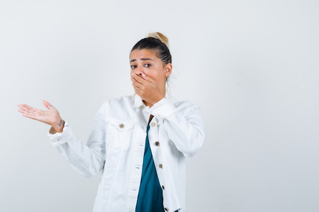 Junge Dame im Hemd, weiße Jacke mit der Hand auf dem Mund, die so tut, als würde sie etwas zeigen und verängstigt aussehen, Vorderansicht.