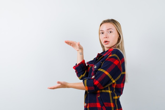 Junge Dame im karierten Hemd, das Größenzeichen zeigt und verwirrt, Vorderansicht schaut.