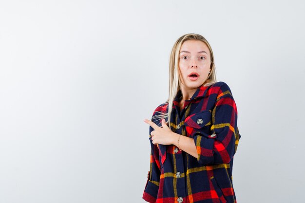 Junge Dame im karierten Hemd zeigt auf die linke Seite und sieht verwirrt aus, Vorderansicht.