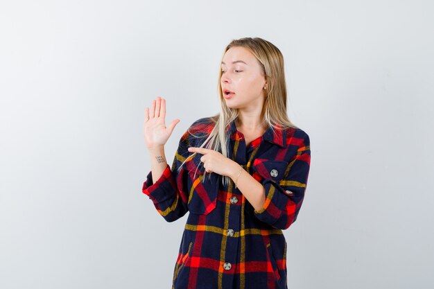 Junge Dame im karierten Hemd zeigt auf Handfläche und schaut erstaunt, Vorderansicht.