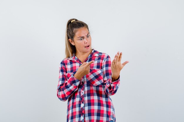 Junge Dame im karierten Hemd zeigt auf ihre leere Handfläche und sieht verwirrt aus, Vorderansicht.