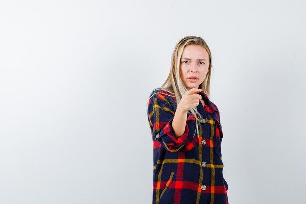 Junge Dame im karierten Hemd zeigt auf Kamera und sieht ernst, Vorderansicht.