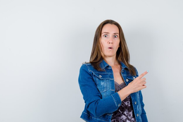 Junge Dame im Kleid, Jeansjacke, die auf die obere rechte Ecke zeigt und verängstigt aussieht, Vorderansicht.