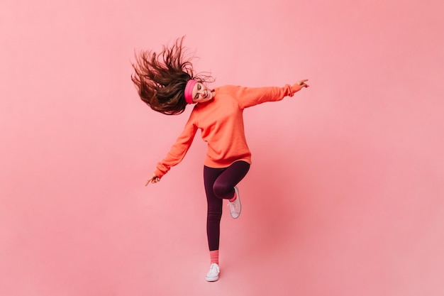 Junge Dame im Sportoutfit tanzt auf rosa Hintergrund Ganzkörperporträt einer Frau in orangefarbenem Sweatshirt