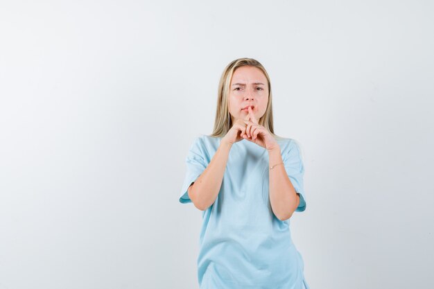 Junge Dame im T-Shirt, das Finger auf Mund hält und ernsthafte Vorderansicht sieht.