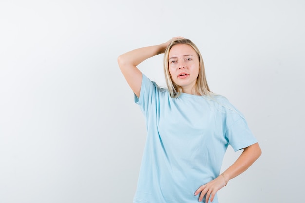 Junge Dame im T-Shirt, das Hand auf Kopf hält, während Hand auf Taille hält und nachdenklich schaut, Vorderansicht.