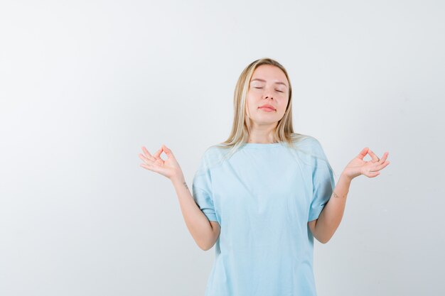 Junge Dame im T-Shirt, das Meditationsgeste zeigt und friedlich, Vorderansicht schaut.