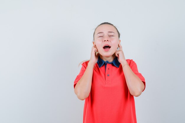 Junge Dame im T-Shirt, das Ohren mit den Fingern verstopft und hoffnungsvoll aussieht