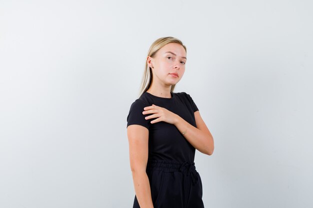 Junge Dame im T-Shirt, Hose, die Hand auf Brust hält und selbstbewusst, Vorderansicht schaut.