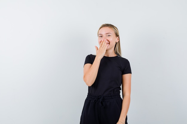 Junge Dame im T-Shirt, Hosen, die Hand auf Mund halten und glücklich schauen, Vorderansicht.