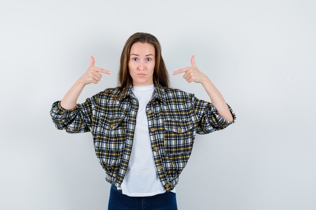 Junge Dame im T-Shirt, in der Jacke, in den Jeans, die auf sich selbst zeigen und stolz schauen, Vorderansicht.