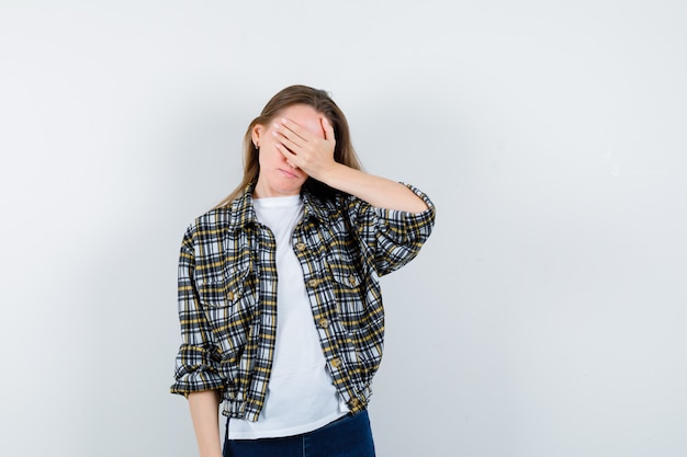 Junge Dame im T-Shirt, in der Jacke, in den Jeans, die Augen mit der Hand bedecken und vergesslich schauen, Vorderansicht.