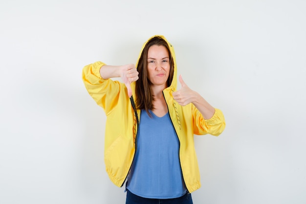 Junge Dame im T-Shirt, Jacke, die Daumen nach oben und unten zeigt und selbstbewusst aussieht, Vorderansicht.