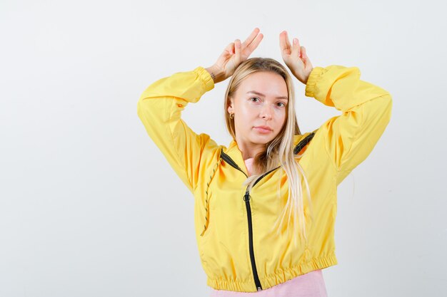 Junge Dame im T-Shirt, Jacke, die Finger über Kopf als Hörner hält und niedlich schaut