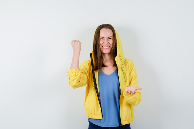 Junge Dame im T-Shirt, Jacke mit Gewinnergeste, Handfläche offen halten und glücklich aussehend, Vorderansicht.