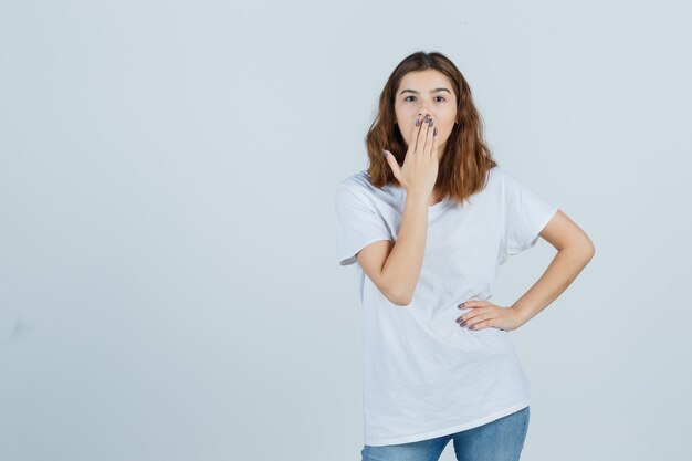 Junge Dame im T-Shirt, Jeans, die Hand auf Mund hält und erstaunt, Vorderansicht schaut.