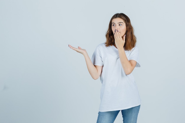 Junge Dame im T-Shirt, Jeans, die Hand auf Mund halten, während sie etwas zeigen und verwirrt schauen, Vorderansicht.